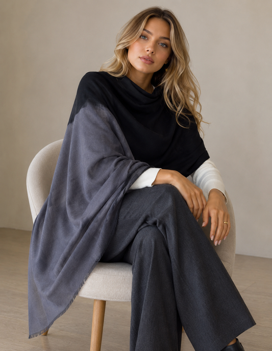 A woman sits on a light beige upholstered chair wearing a large draped shawl in black and dark gray, layered over a white long-sleeve top and dark wide-leg pants, against a neutral beige wall.