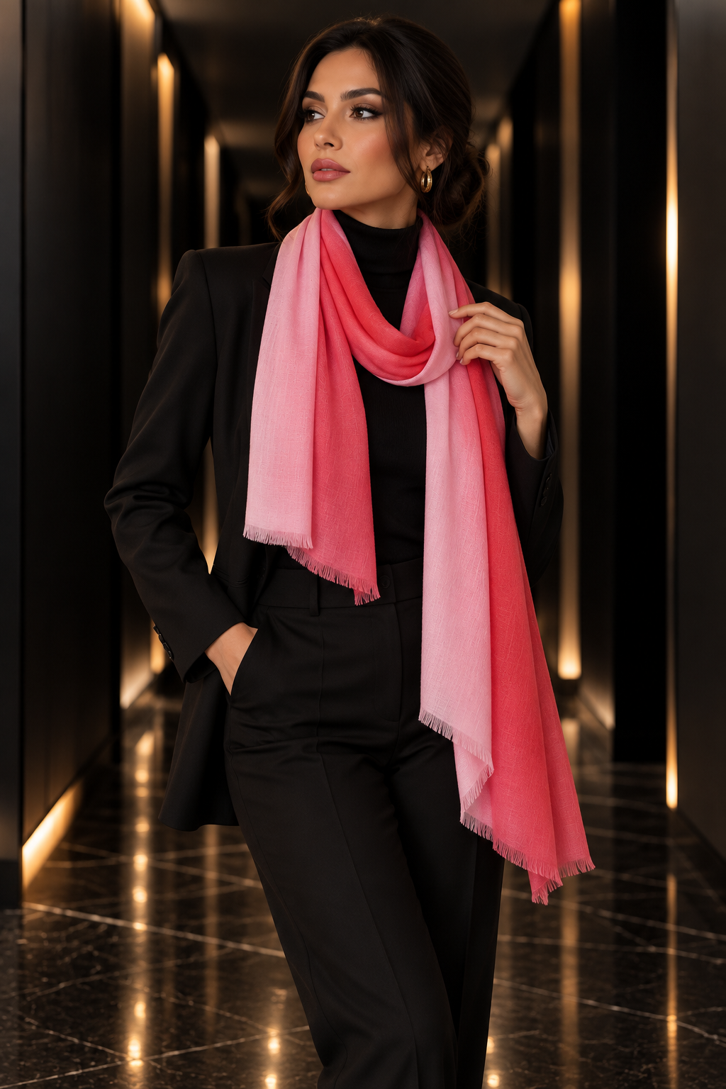 A woman wearing a black suit and a pink gradient scarf with fringed edges, standing in a modern hallway with warm lighting.