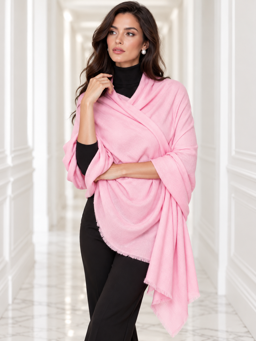 A woman modeling a light pink shawl draped over her shoulders, paired with a black turtleneck and black pants, standing in a bright white hallway with classical wall moldings.