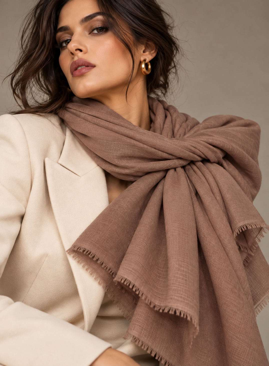 A woman wearing a large, soft taupe-brown scarf tied around her neck, styled with a cream blazer and gold hoop earrings against a neutral gray background.