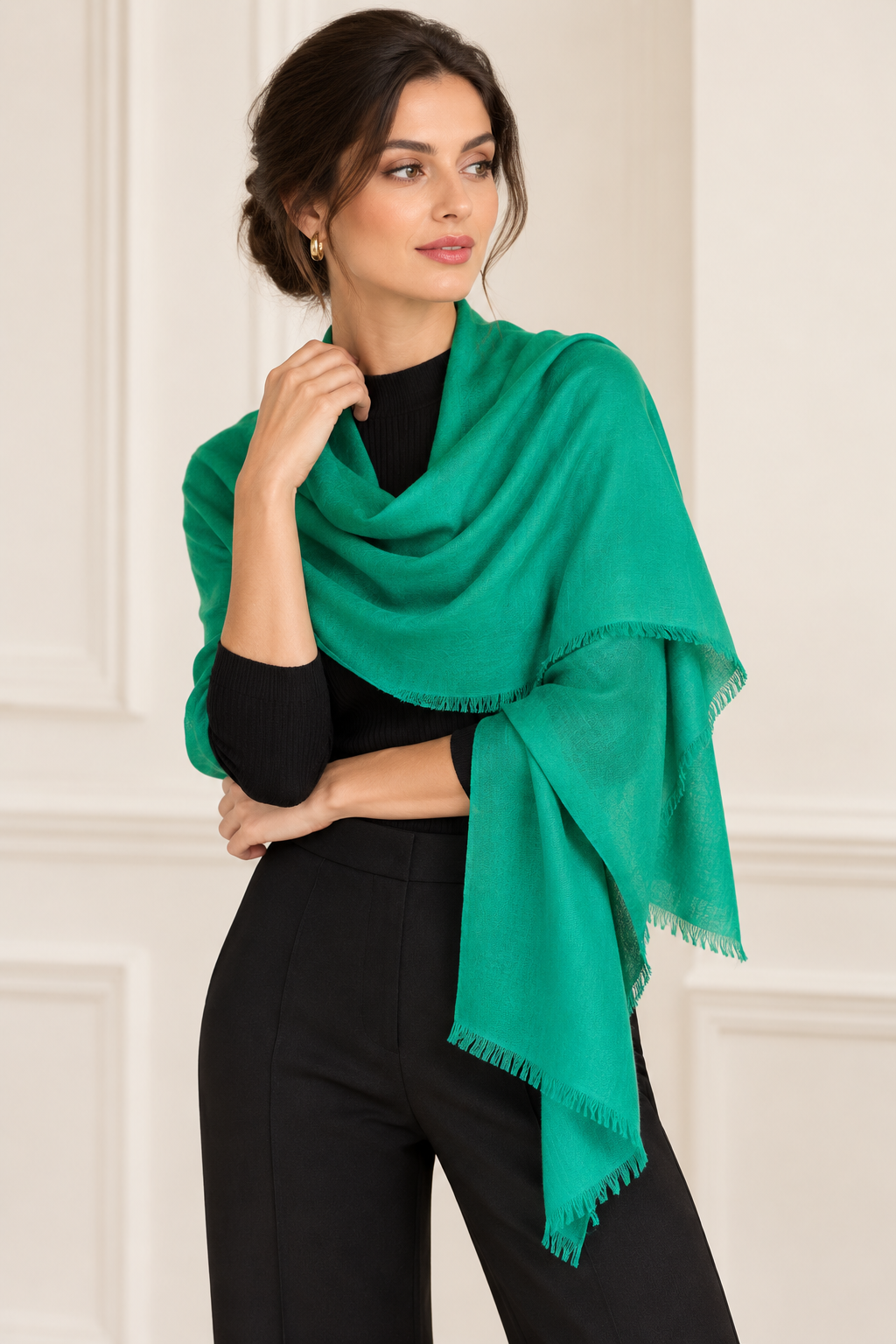 A woman wearing a bright green shawl wrap with fringed edges over a black top and black pants, standing against a light cream paneled wall.