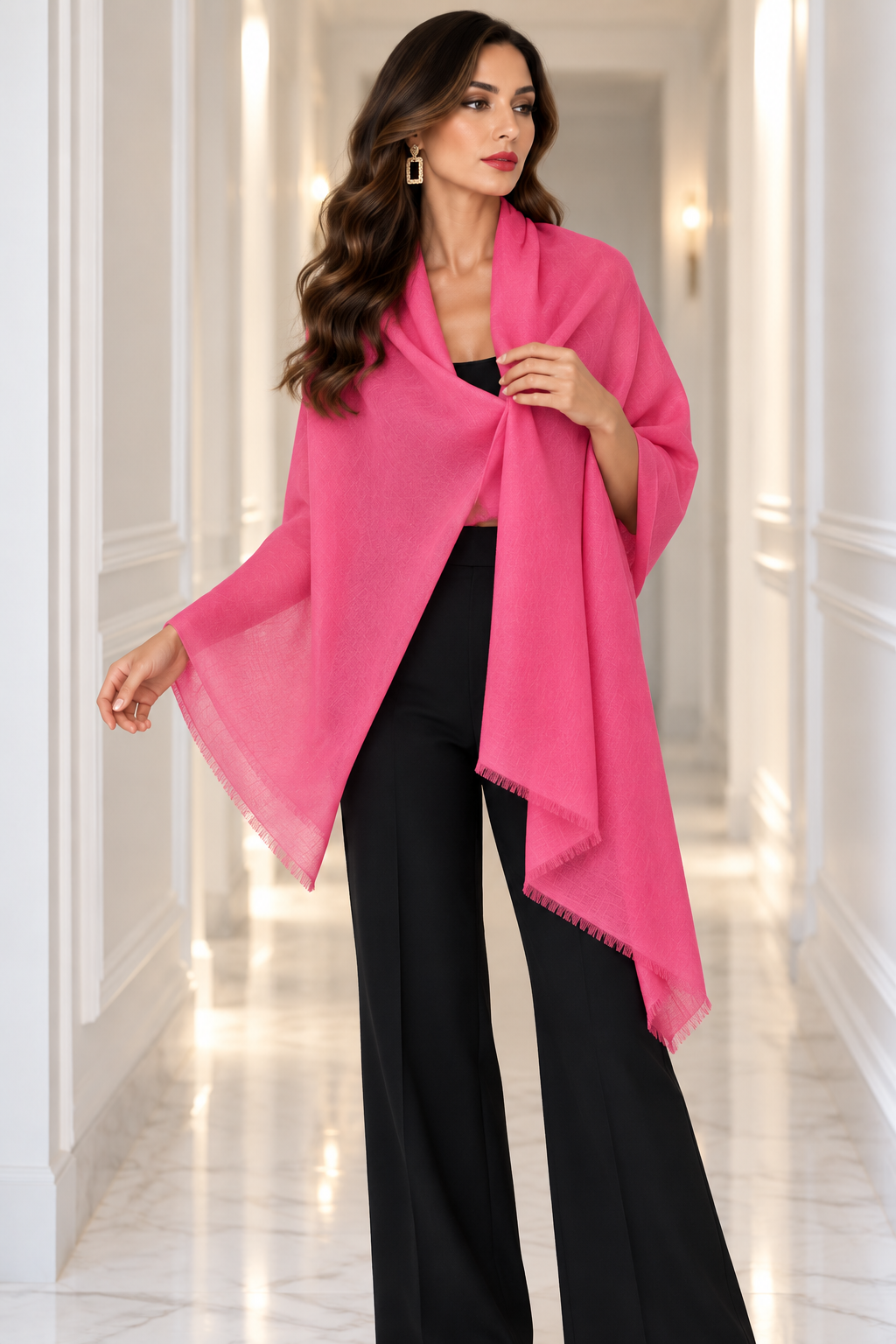 A woman modeling a bright pink lightweight shawl or wrap draped over her shoulders, paired with black wide-leg pants, in a bright white hallway.