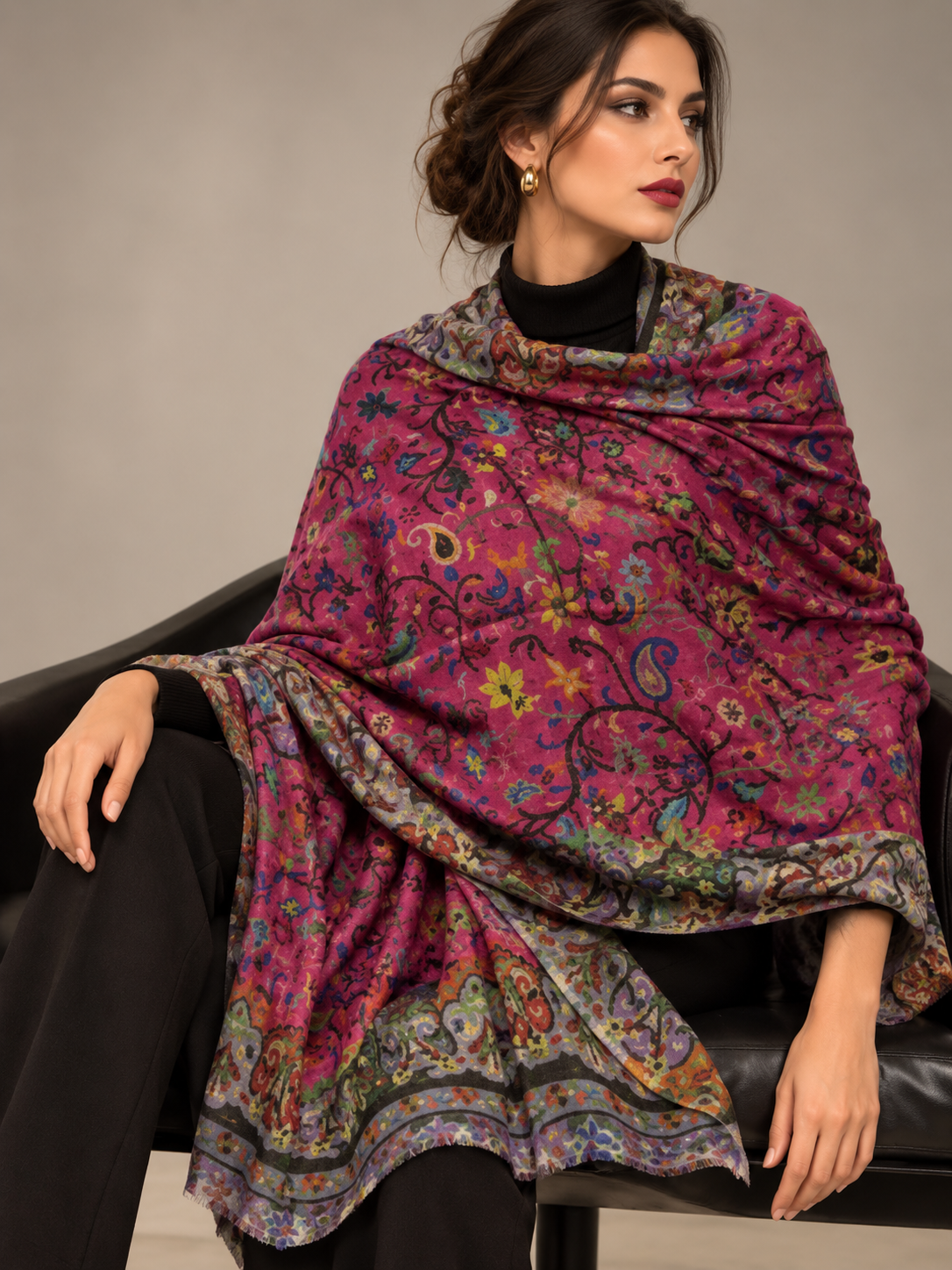 A woman seated on a black leather chair wearing a large, draped shawl with a vibrant magenta base and intricate multicolor paisley and floral patterns.