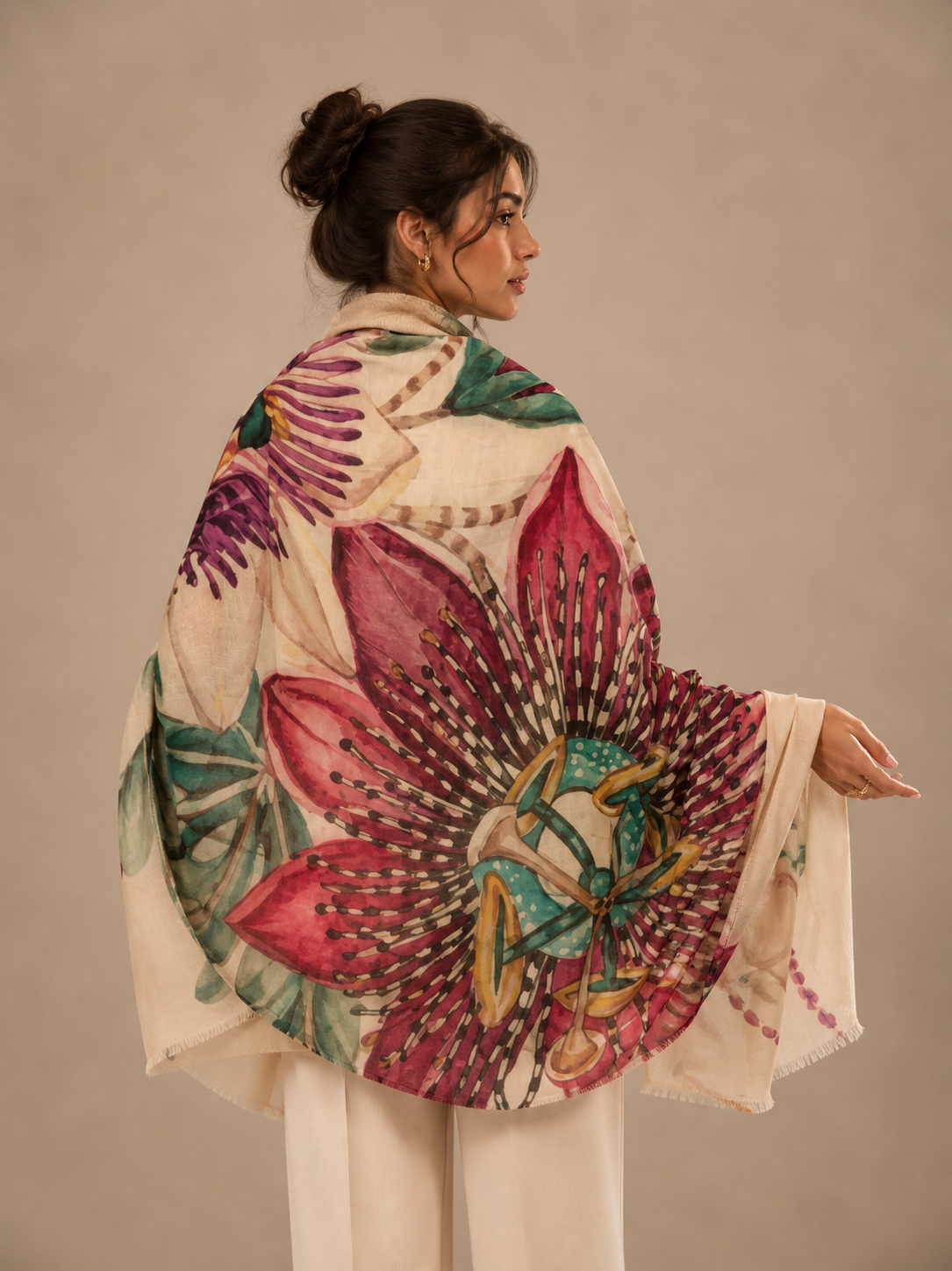 A woman shown from the back wearing a large cream shawl or wrap with a bold floral print. The print features a prominent magenta-pink flower with green leaves and purple accents, styled with cream trousers against a solid beige studio background.