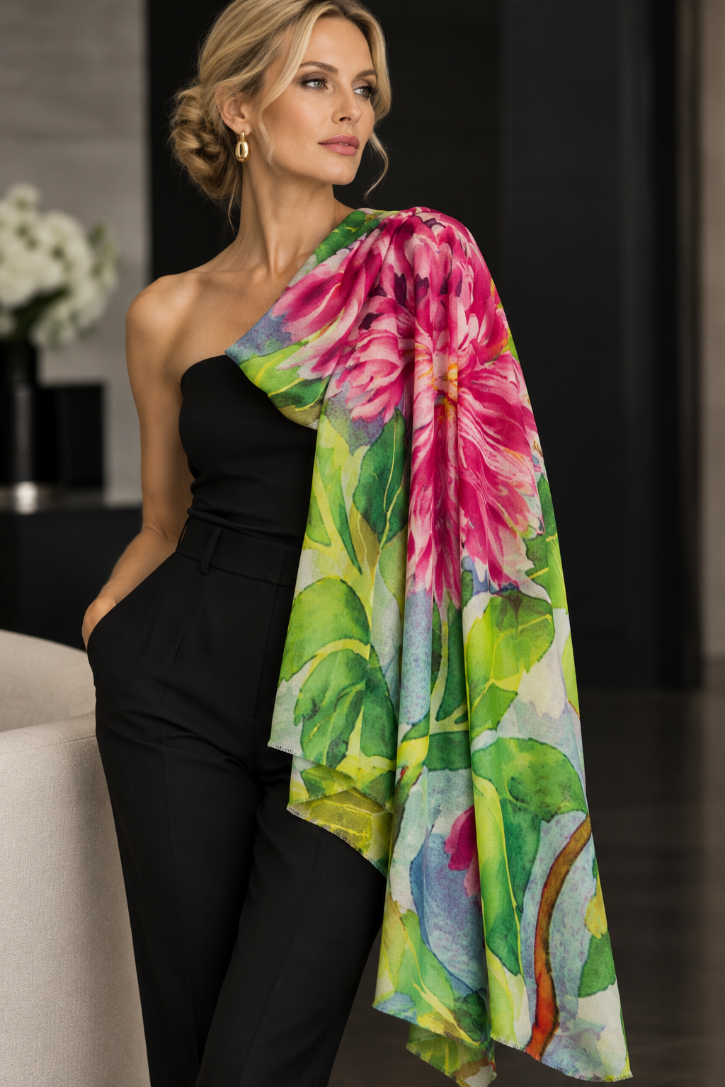 A woman wearing a black strapless jumpsuit and gold hoop earrings models a vibrant floral shawl draped over her shoulder. The shawl features a large pink flower and green leaves in a watercolor print, set against a dark, elegant indoor background.