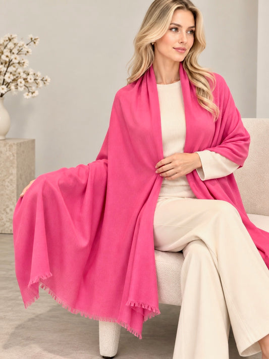 A woman seated on a light upholstered chair wearing a bright pink shawl draped over her shoulders, paired with a cream top and cream wide-leg pants, in a softly lit indoor setting.