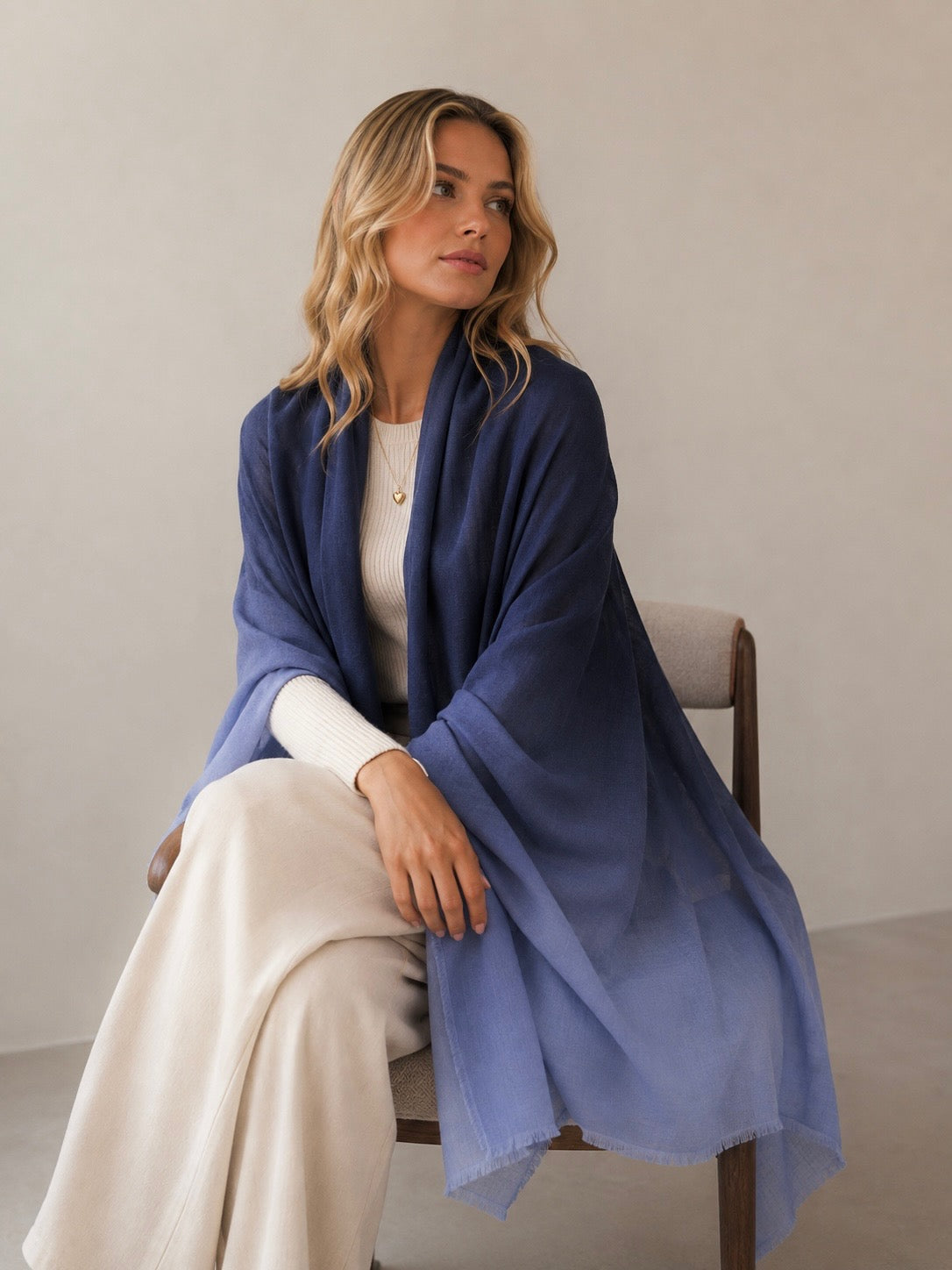A woman sits on a wooden chair wearing a large navy-to-light-blue ombre shawl draped over her shoulders, paired with a cream top and cream wide-leg pants, against a neutral beige wall.