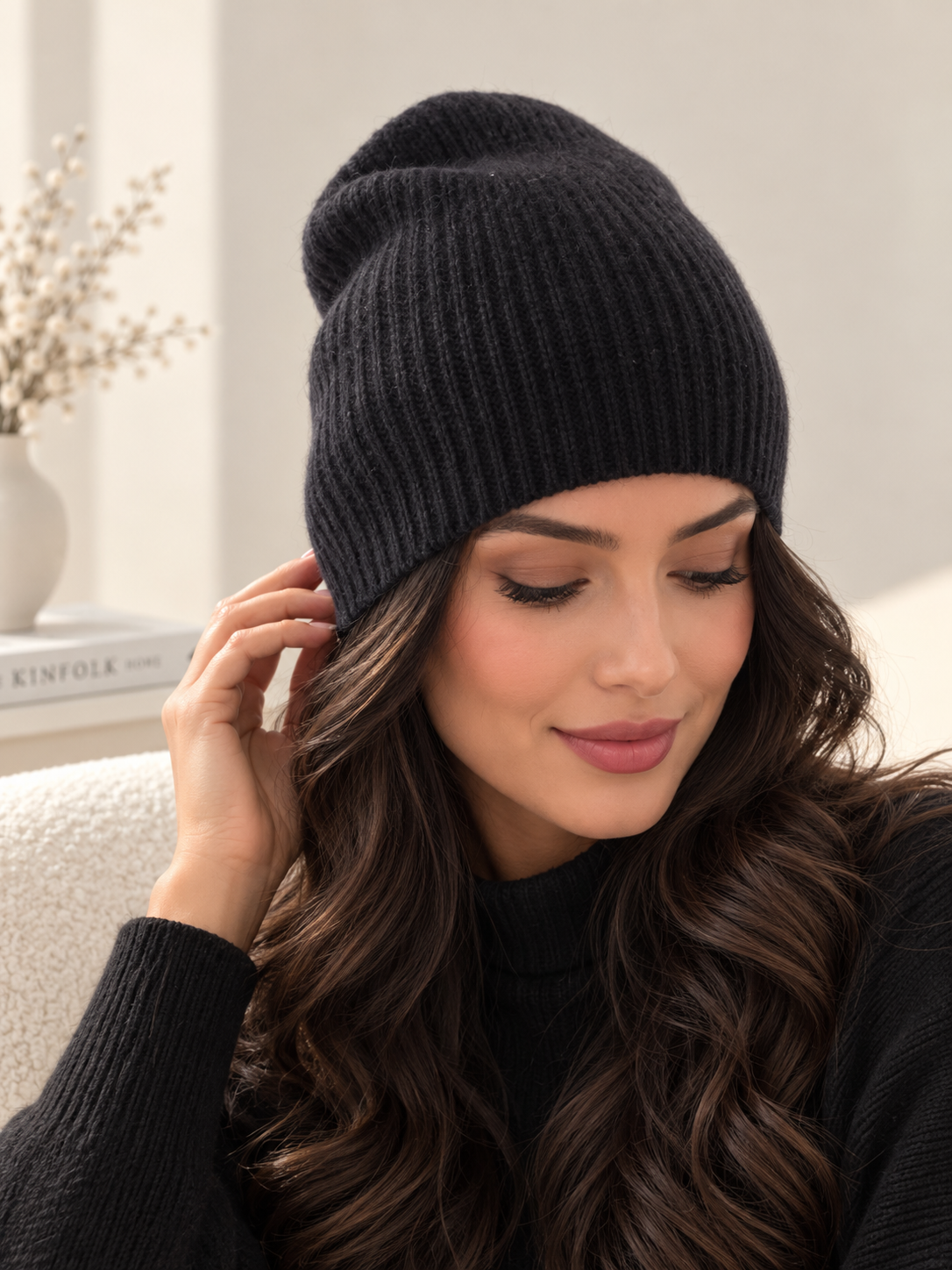 A woman wearing a black ribbed knit beanie hat, shown in a warm indoor setting with soft light and a neutral background.