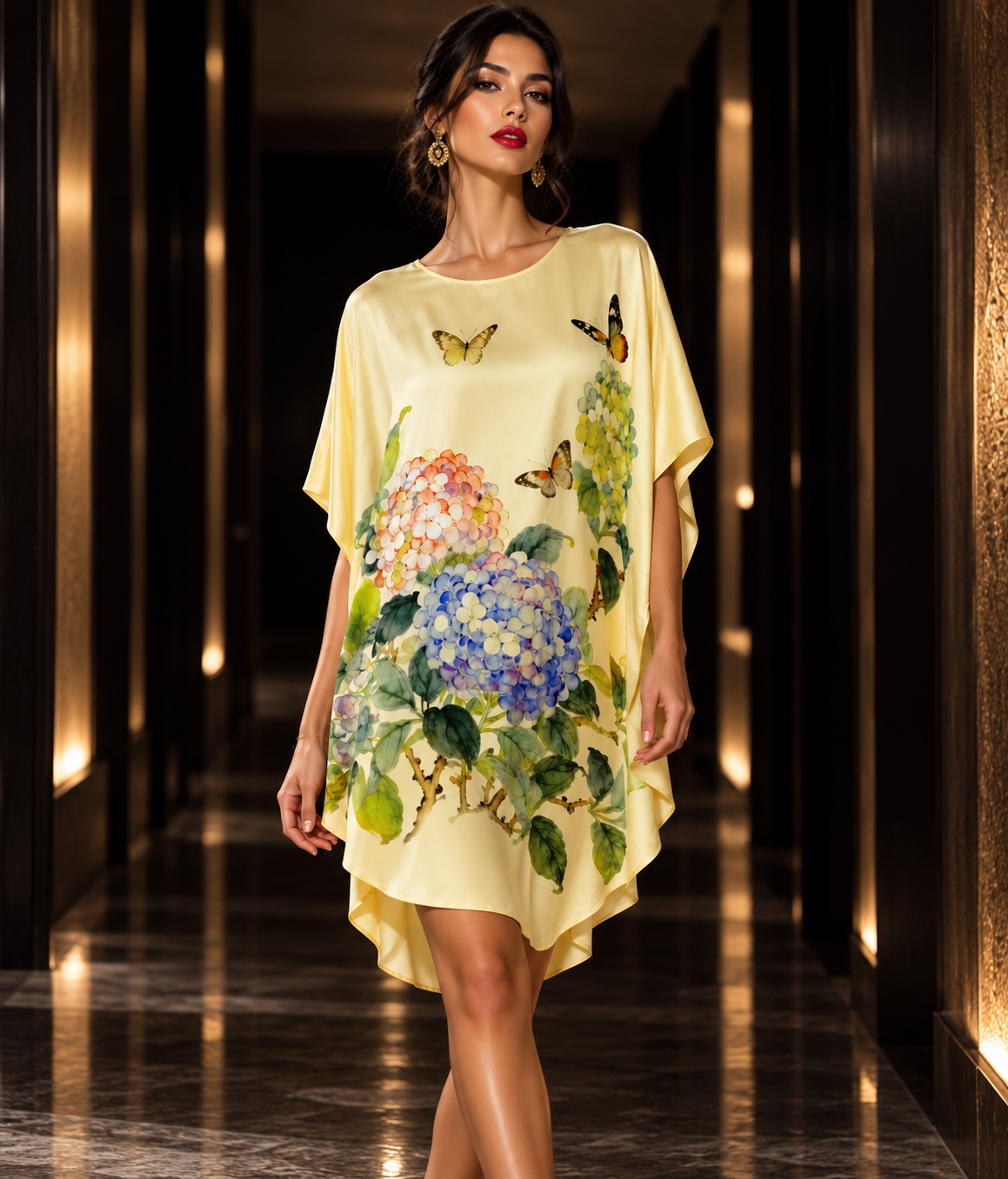 A woman models a pale yellow high-low dress with a watercolor print of hydrangeas and butterflies in a hallway with warm lighting.