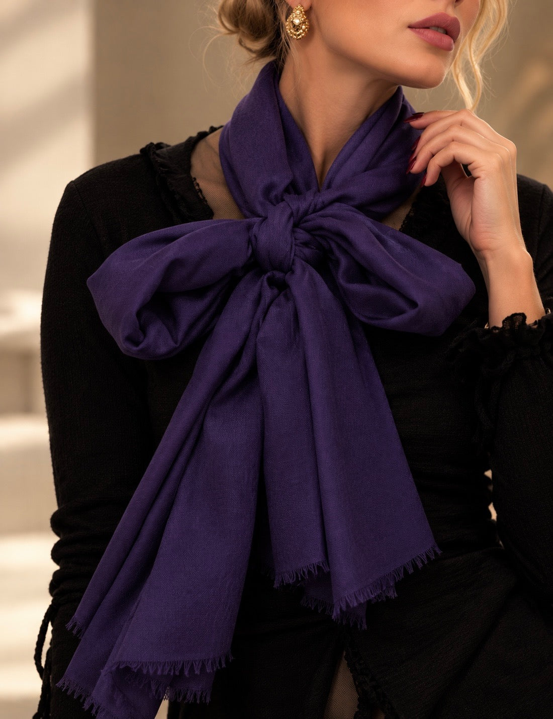 A woman wearing a large purple scarf tied in a bow around her neck, paired with a black top. The photo is cropped from the mouth down, showing the scarf's drape and texture in warm indoor lighting.
