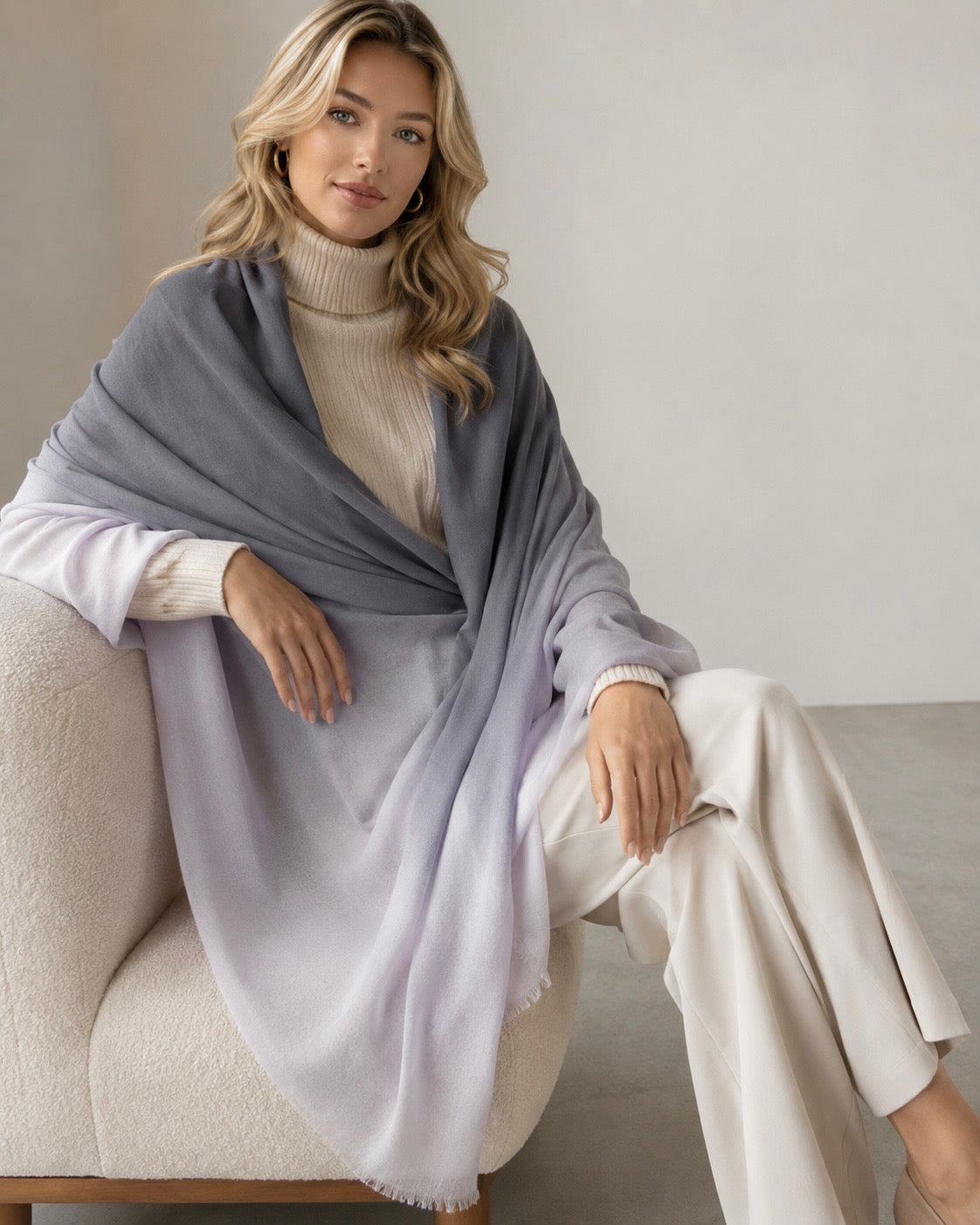 A woman seated on a cream upholstered chair wearing a large gradient shawl or pashmina draped over her shoulders, paired with a cream turtleneck and cream wide-leg pants in a softly lit indoor setting.