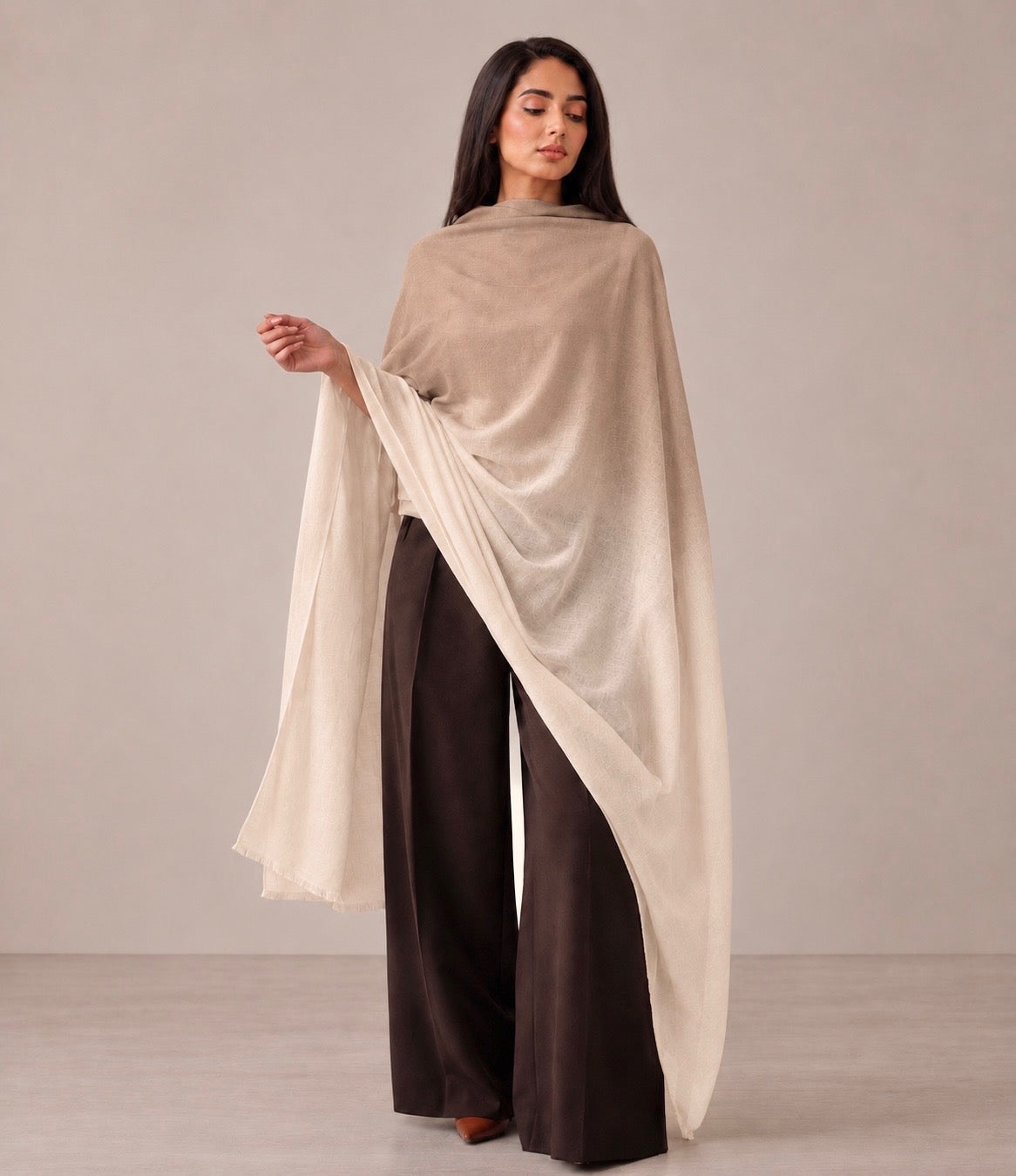 A woman models a lightweight draped shawl in a warm beige color, worn over dark brown wide-leg trousers against a neutral studio backdrop.