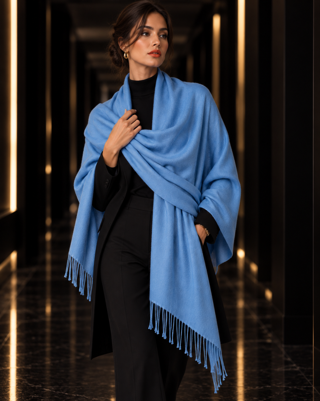 A woman wearing a light blue fringed shawl over a black outfit, standing in a dark, elegant hallway with warm vertical lights.