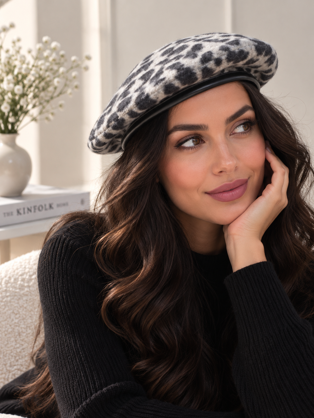 A woman wearing a leopard-print beret and a black ribbed sweater, resting her chin on her hand in a softly lit indoor setting.
