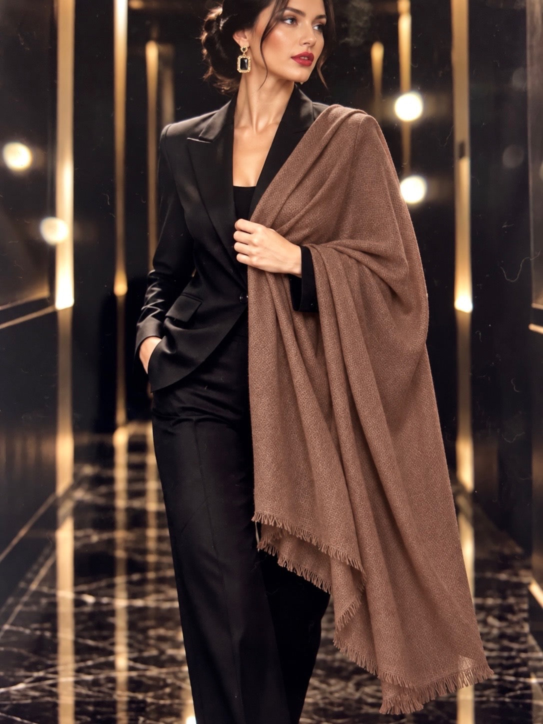 A woman models a luxurious brown shawl with fringed edges, draped over her shoulders and held at her waist, paired with a black blazer and trousers in a modern hallway with gold-toned lighting.