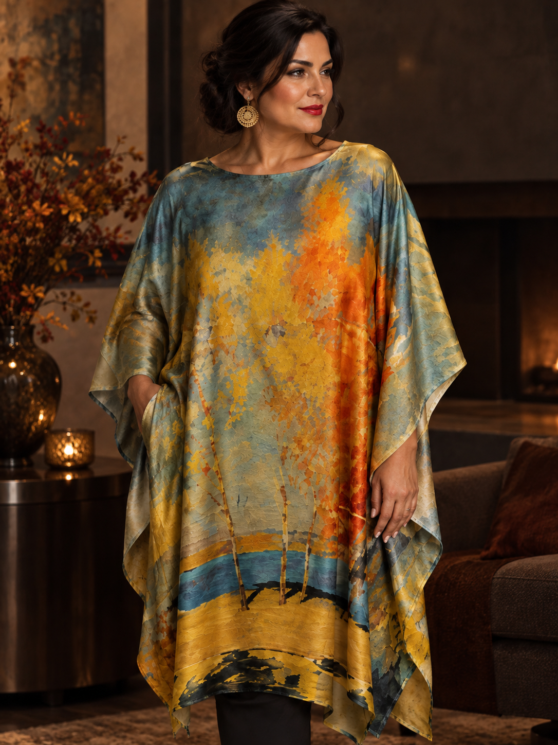 A woman wearing a loose, flowy kaftan-style top with an abstract painterly print in blue, yellow, orange, and beige tones, posed in a warm, dimly lit living room interior.