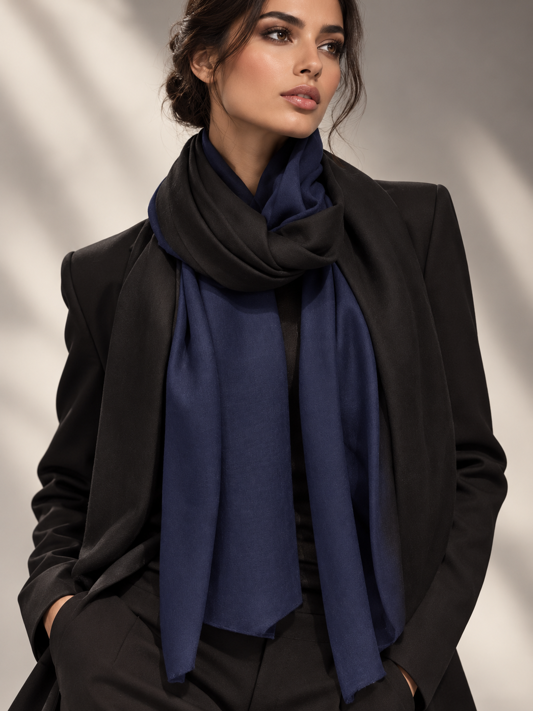 A woman wearing a black blazer and trousers with a navy blue scarf draped around her neck, photographed against a softly lit neutral background with leaf shadows.