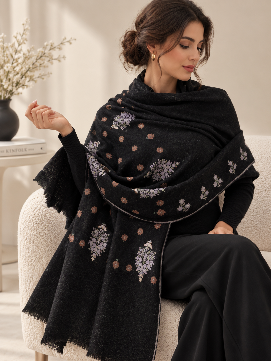 A woman seated on a cream upholstered chair wearing a black shawl with embroidered floral motifs in lavender, beige, and muted brown. The setting is a softly lit indoor room with a side table and dried flowers in the background.