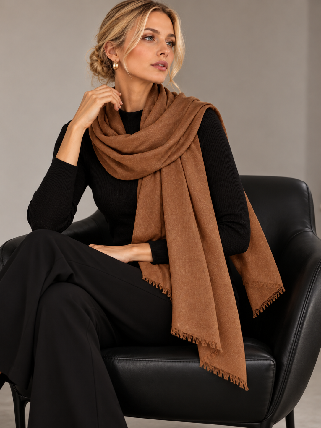 A woman seated in a black leather chair wearing a black outfit and a large camel-brown scarf with fringed edges, photographed against a plain light gray studio background.
