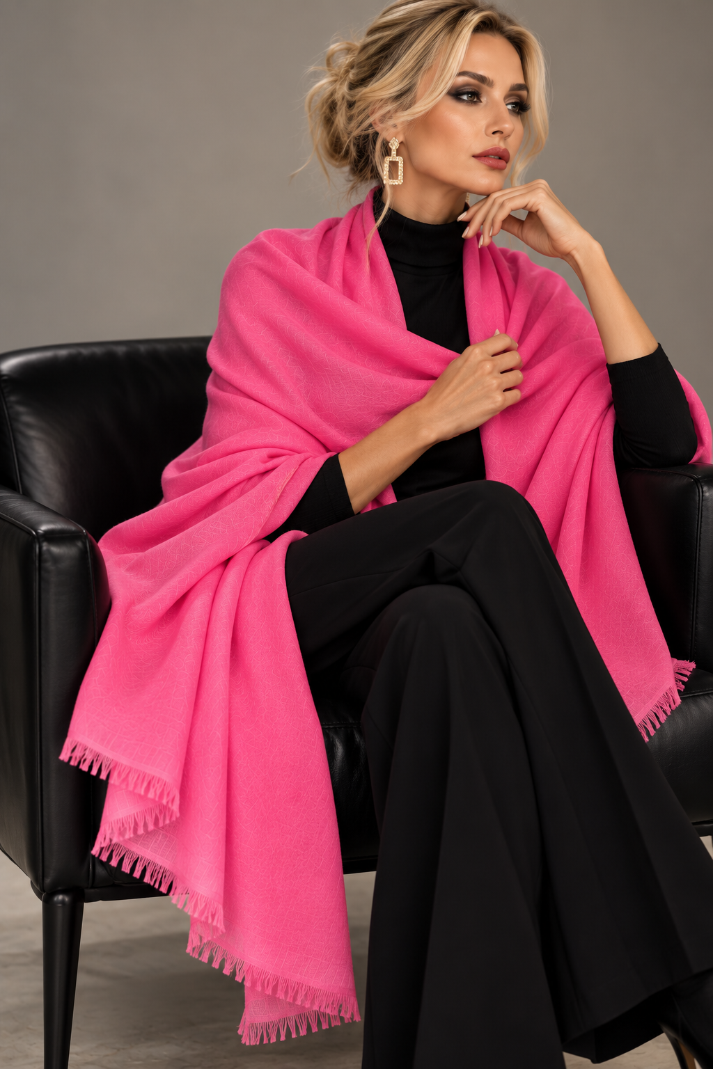 A woman seated in a black leather chair wearing a bright pink shawl over a black outfit, with gold geometric earrings, against a plain gray studio background.