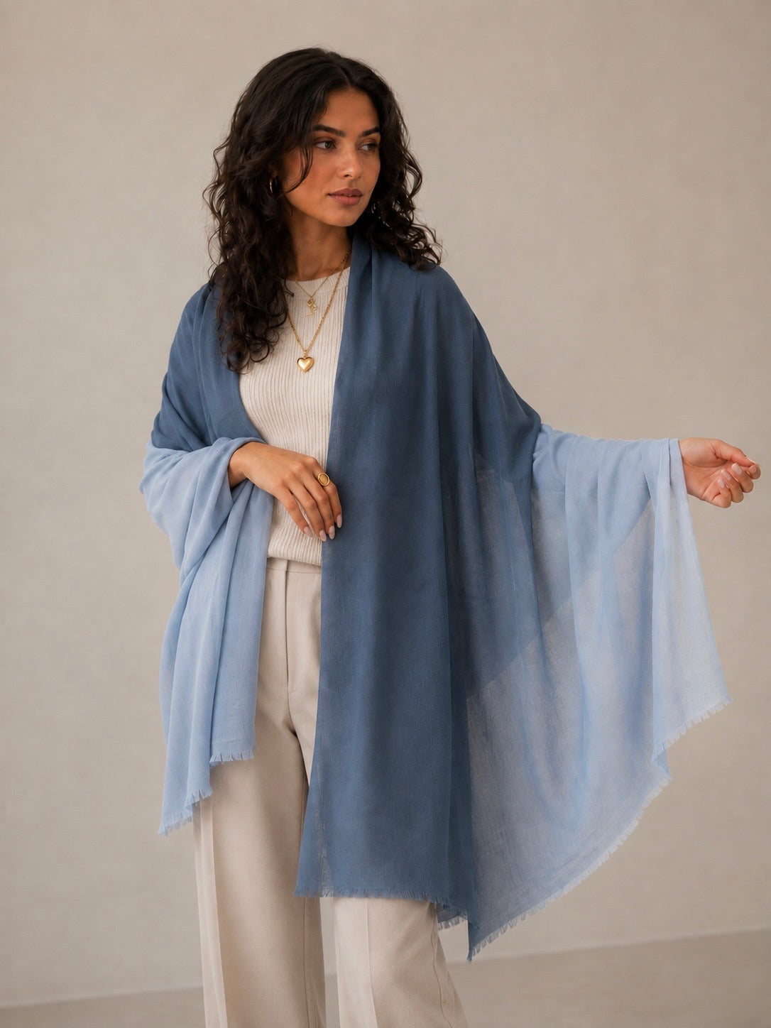 A woman modeling a lightweight blue shawl draped over her shoulders, worn over a cream ribbed top and cream trousers, against a neutral beige studio background.