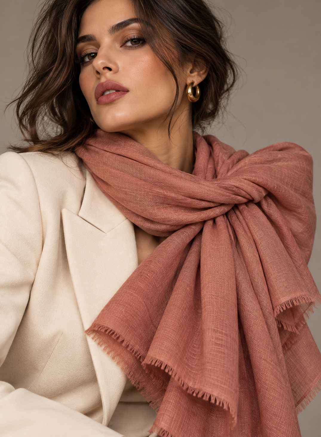 A woman wearing a dusty rose pink shawl scarf with fringed edges, styled over a cream blazer. She is wearing gold hoop earrings against a neutral gray background.