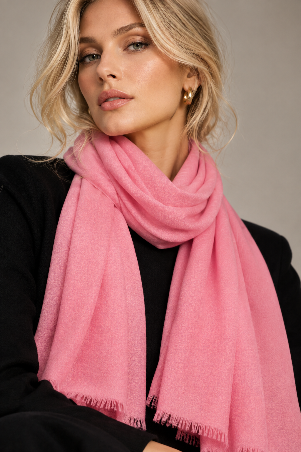 A woman with blonde hair wearing a soft pink scarf and a black top, photographed against a neutral gray studio background.
