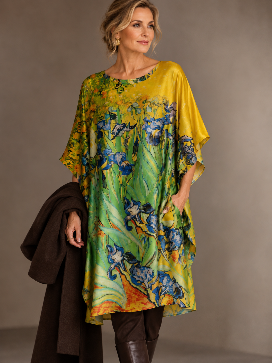 A woman models a loose, short-sleeved tunic top with a vibrant yellow and green base featuring a blue and white floral pattern, styled with a dark brown coat and brown pants against a neutral studio backdrop.