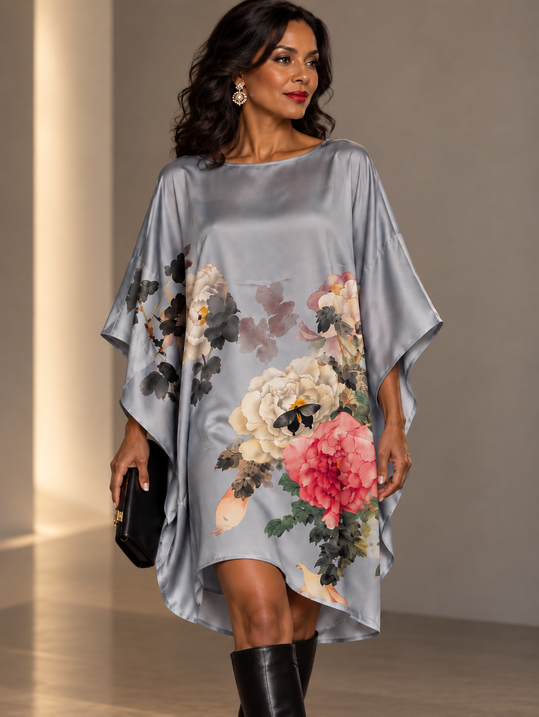 A woman models a silk kaftan dress featuring a large floral print with pink, white, and green peonies. She is wearing black leather boots and holding a black clutch bag in a softly lit indoor setting.
