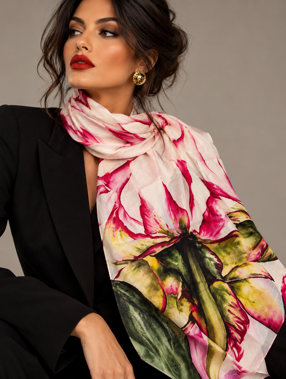 A woman wearing a large floral scarf with pink, green, and yellow tones, styled over a black blazer against a neutral gray studio background.