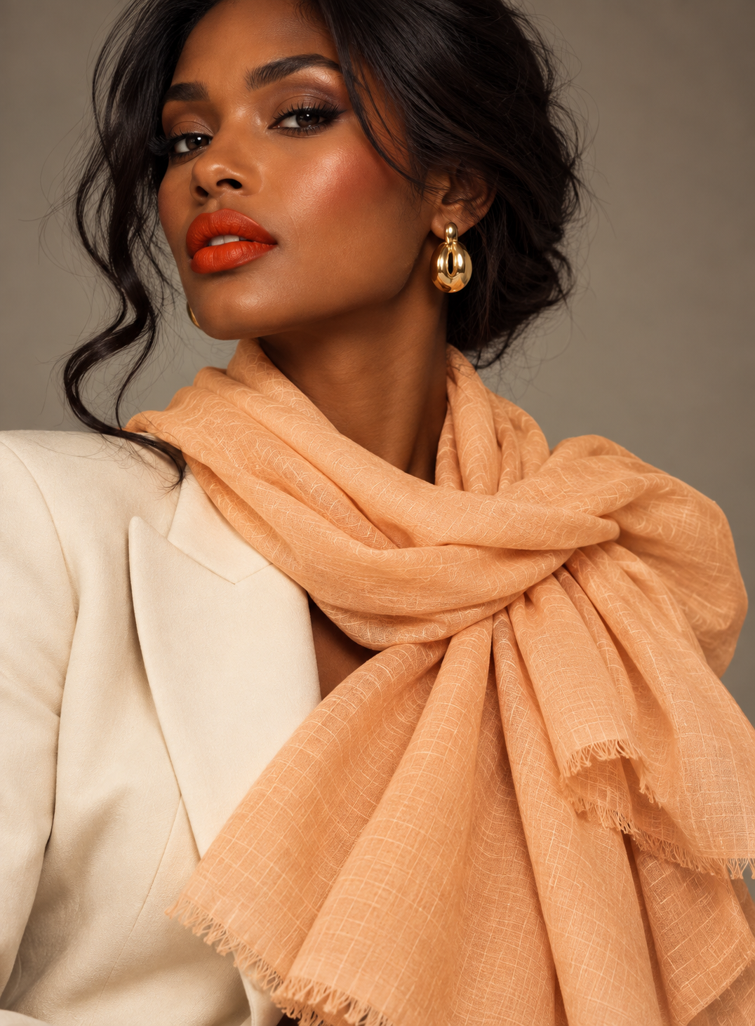 A woman wearing a peach-colored lightweight scarf with fringed edges, styled over a cream blazer. She is wearing gold teardrop earrings against a neutral gray background.