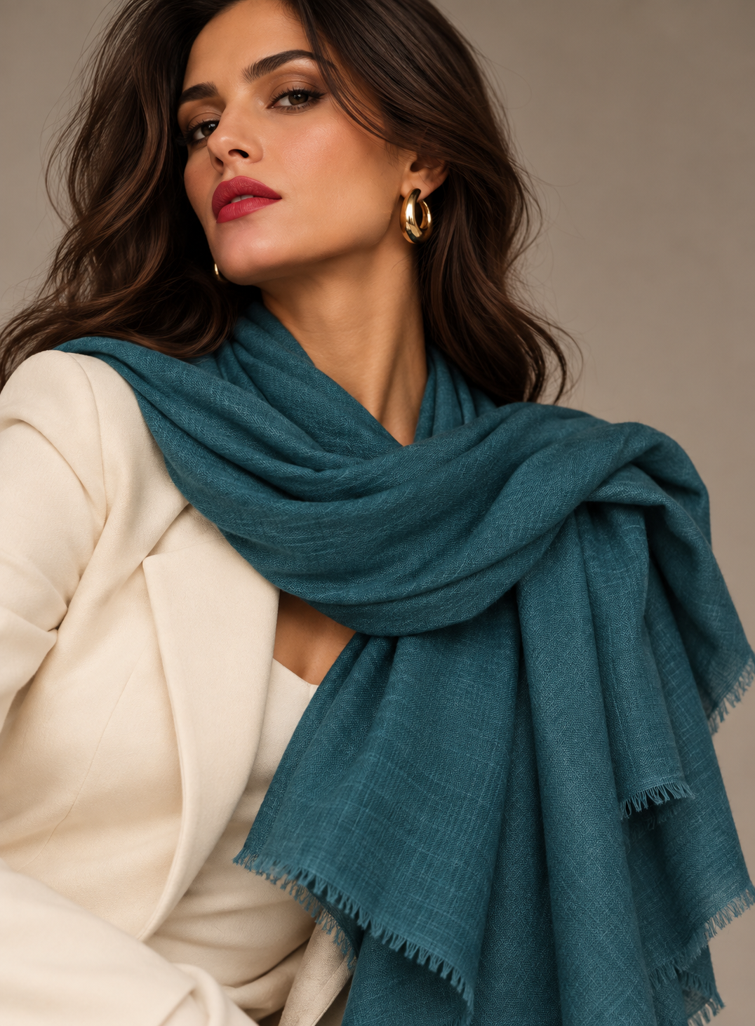 A woman wearing a teal blue-green lightweight scarf with fringed edges over a cream blazer, set against a neutral beige background.