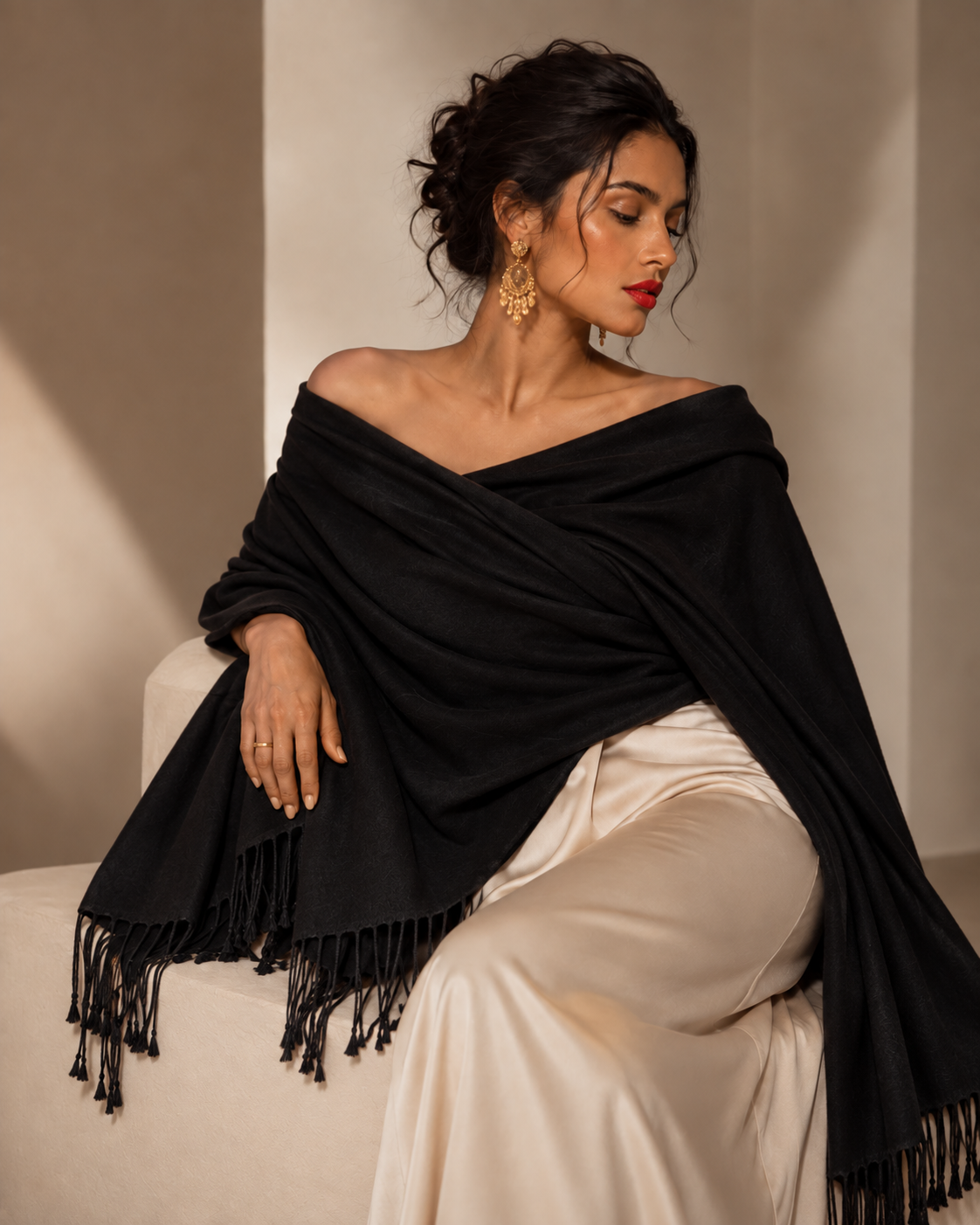 A woman seated indoors wearing a black off-the-shoulder shawl with fringe trim over cream satin pants. She is styled with gold earrings, and the background is a warm beige wall with soft shadows.