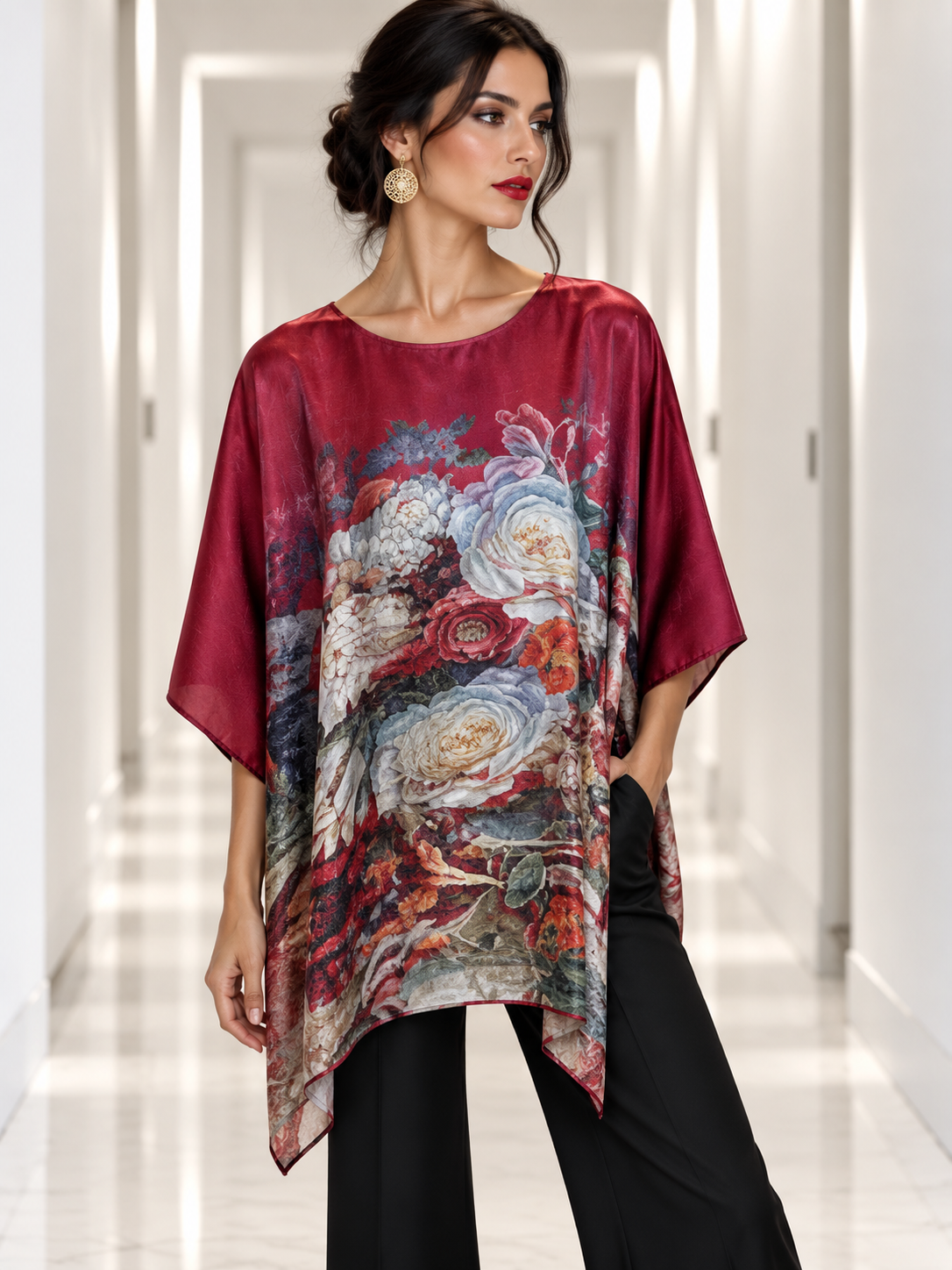 A woman models a loose, short-sleeve top with a deep burgundy base and a large, detailed floral print in red, white, and blue. She is wearing black pants and gold earrings, standing in a brightly lit hallway.