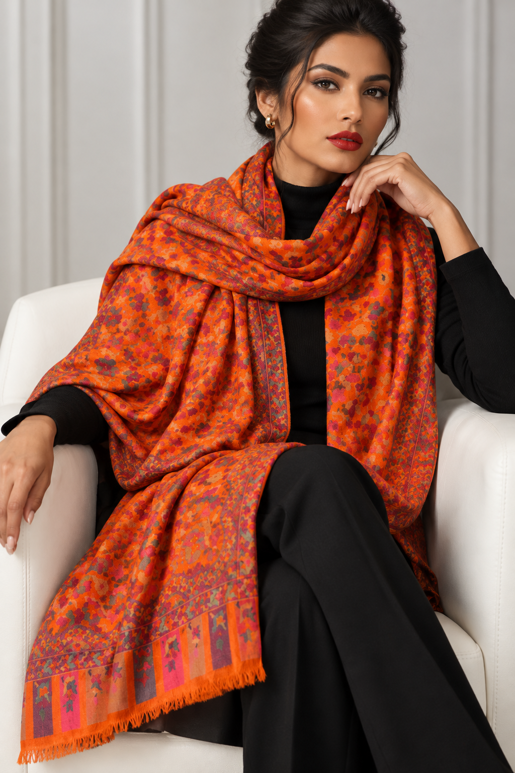 A woman seated in a white armchair wearing a black outfit and a large orange shawl with a multicolor floral and tree pattern, posed against a light gray paneled wall.