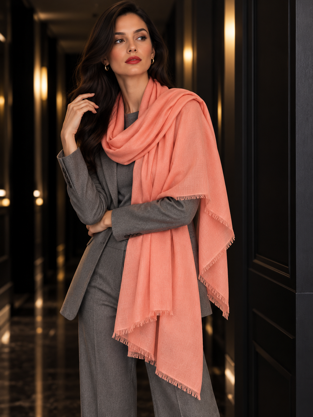 A woman wearing a gray suit and a large peach-pink fringed scarf, standing in a dark, elegant hallway with warm lights.