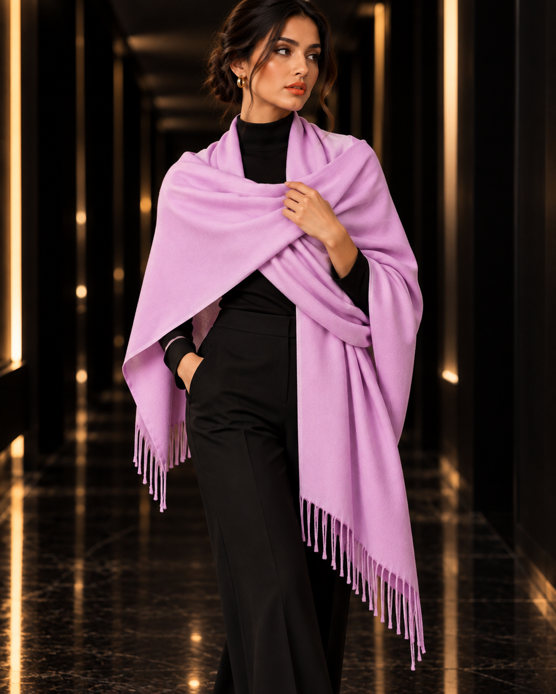A woman models a light purple/lavender pashmina shawl with fringe trim, draped over her shoulders and arms, paired with a black turtleneck and black trousers, set against a dark, elegant hallway with warm lighting.