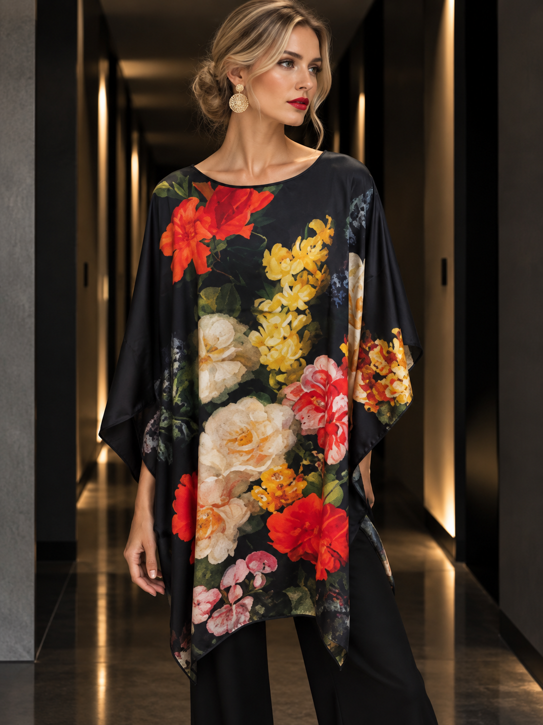 A woman wearing a loose black tunic dress with a large multicolor floral print, standing in a modern hallway with warm lighting.