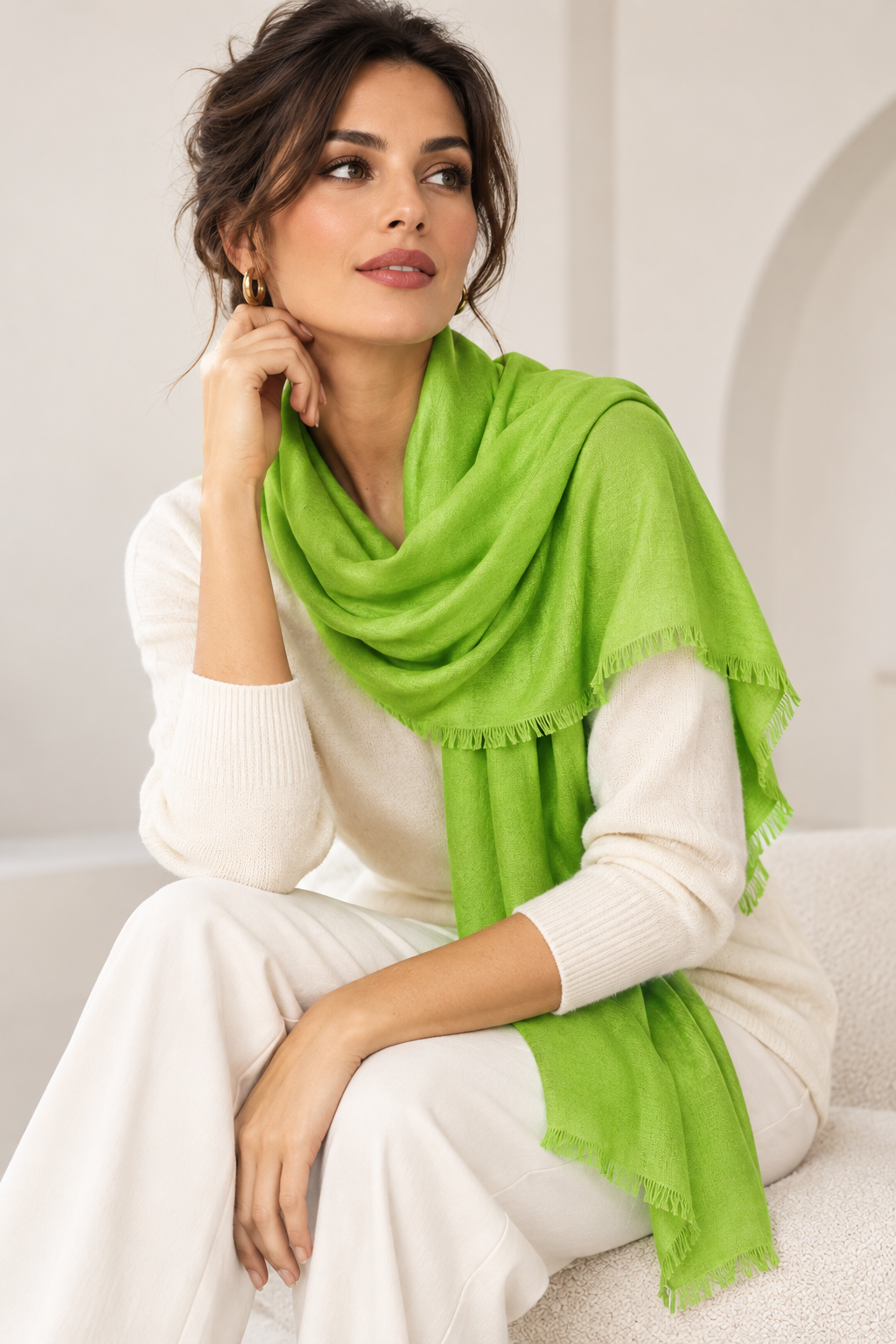 A woman wearing a white ribbed knit sweater and matching pants, draped with a bright lime green shawl scarf featuring fringed edges, sitting in a softly lit indoor setting.