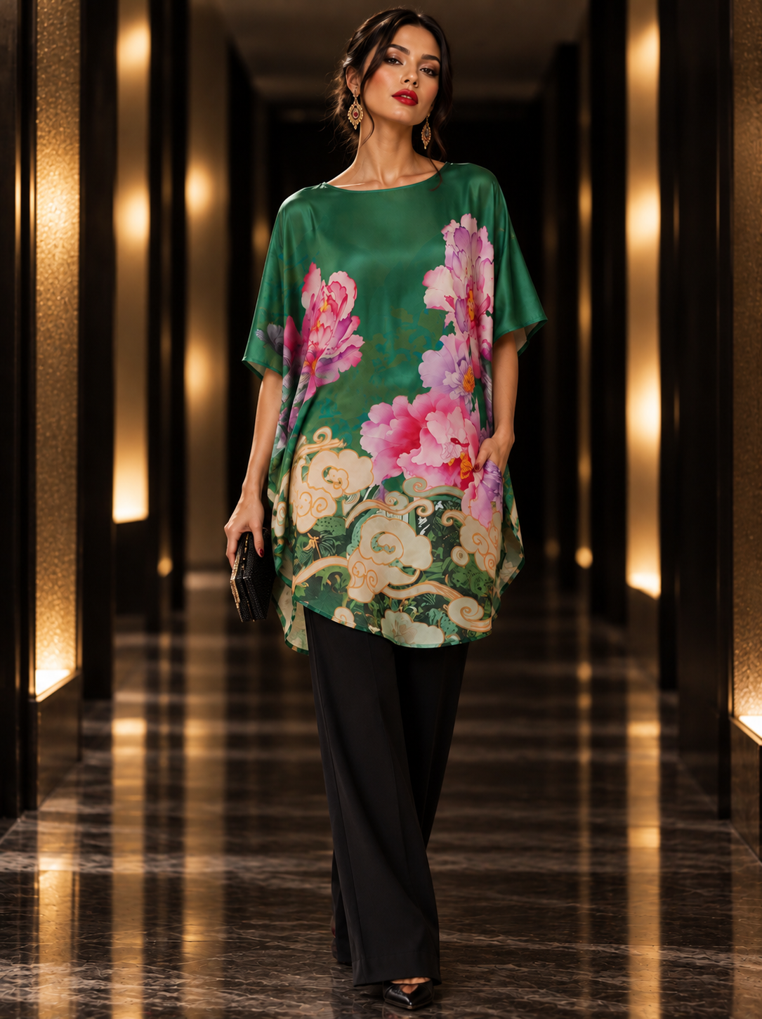 A woman walks down a luxury hallway wearing a green short-sleeve tunic top with a large pink and green floral print, paired with black wide-leg pants.